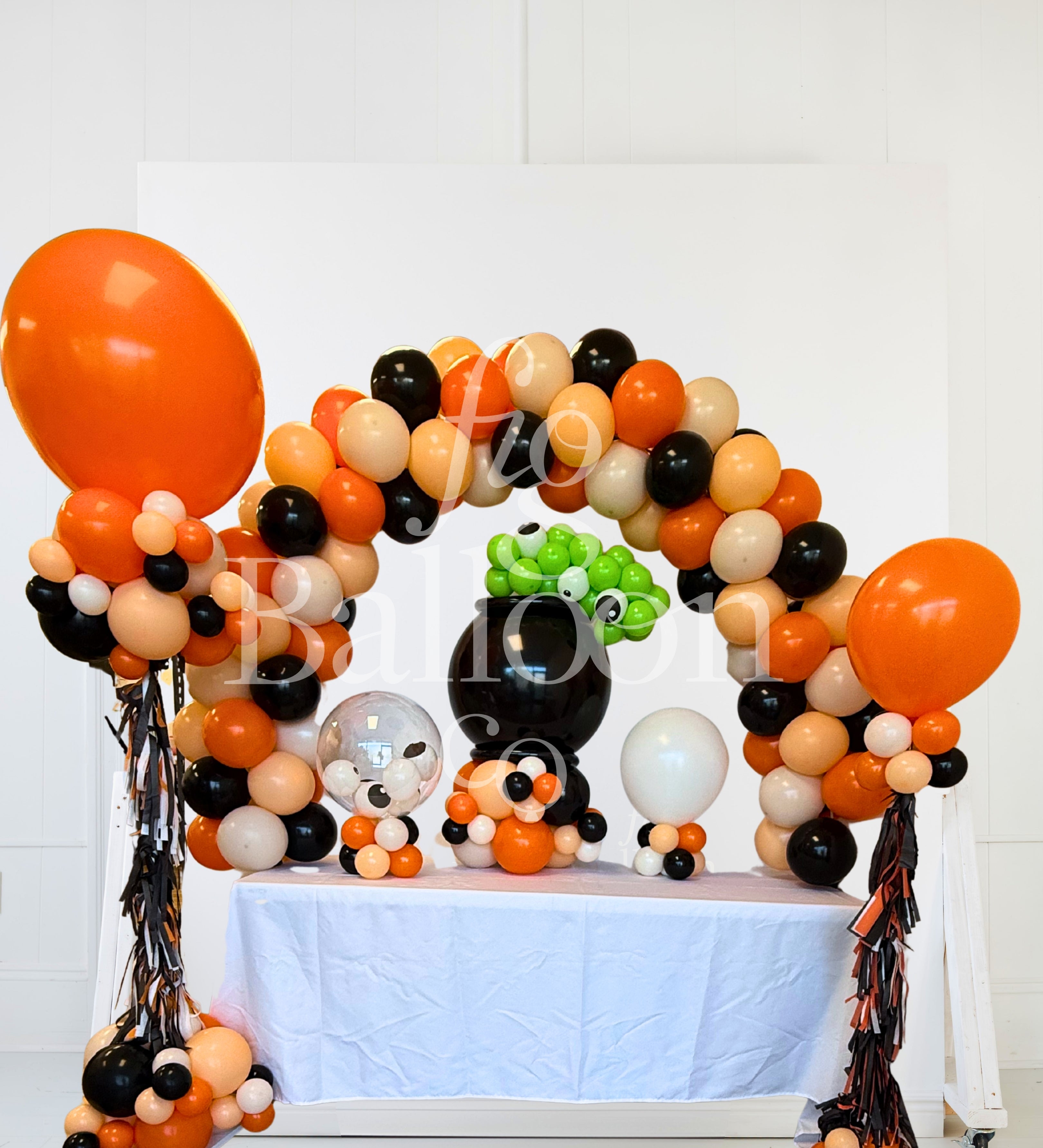 Halloween party balloon decoration setup featuring an orange, black, and beige balloon arch, a large orange balloon, a black pumpkin balloon, a green leaf balloon, and tinsel streamers on a white table runner.