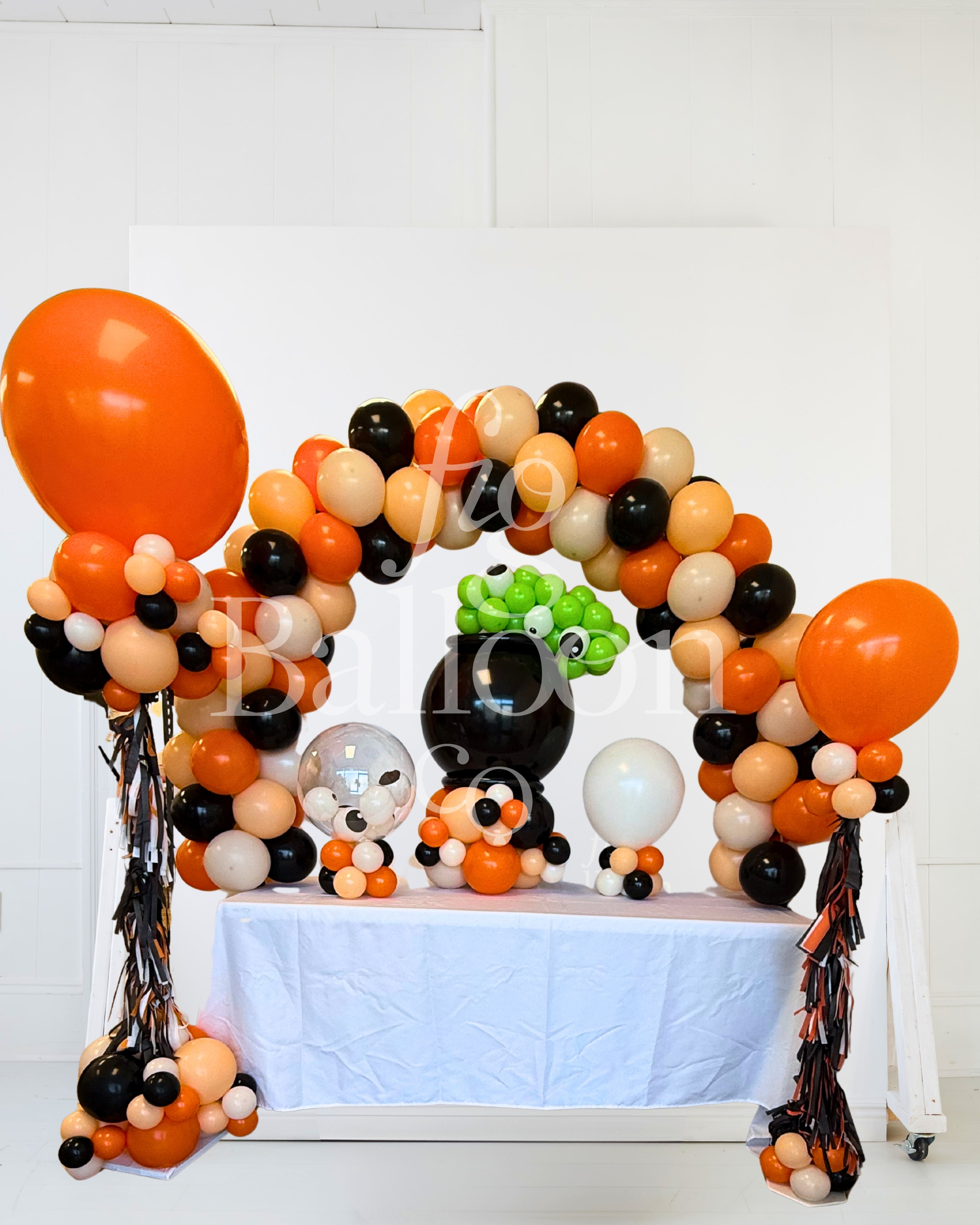 Halloween party balloon decoration setup featuring an orange, black, and beige balloon arch, a large orange balloon, a black pumpkin balloon, a green leaf balloon, and tinsel streamers on a white table runner.