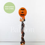 A Halloween-themed balloon bouquet featuring a large orange balloon with 'TRICK OR TREAT' in black text, smaller orange, white, and black balloons, and a long black, orange, and white tassel. A green circle in the top left says 'Add custom text! (+$15)'.