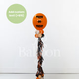 A Halloween-themed balloon bouquet featuring a large orange balloon with 'TRICK OR TREAT' in black text, smaller orange, white, and black balloons, and a long black, orange, and white tassel. A green circle in the top left says 'Add custom text! (+$15)'.