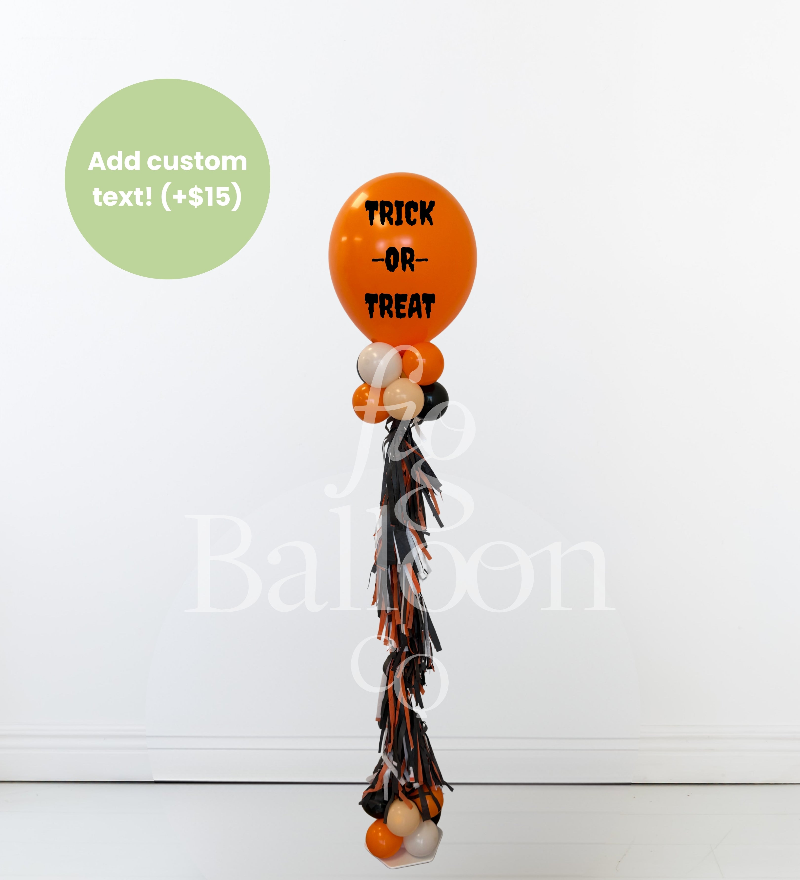 A Halloween-themed balloon bouquet featuring a large orange balloon with 'TRICK OR TREAT' in black text, smaller orange, white, and black balloons, and a long black, orange, and white tassel. A green circle in the top left says 'Add custom text! (+$15)'.