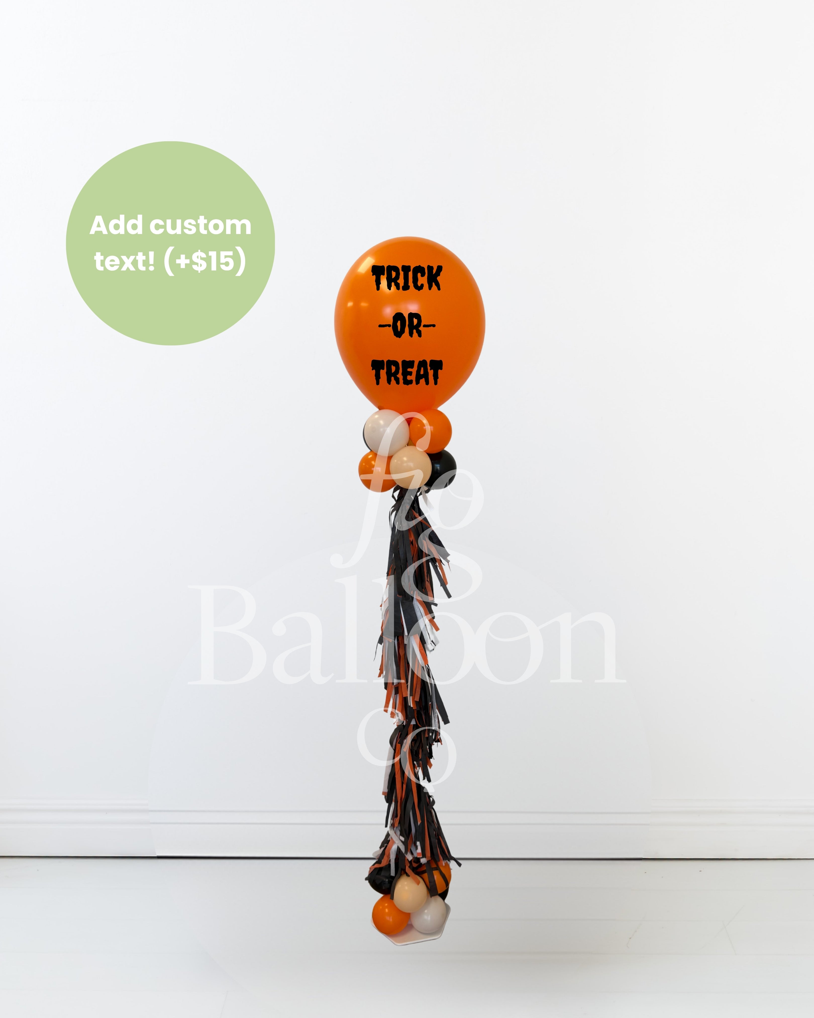 A Halloween-themed balloon bouquet featuring a large orange balloon with 'TRICK OR TREAT' in black text, smaller orange, white, and black balloons, and a long black, orange, and white tassel. A green circle in the top left says 'Add custom text! (+$15)'.