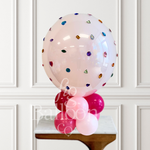 A large clear pink balloon decorated with scattered colorful gem-like confetti, displayed on top of a cluster of pink and light pink balloons on a small table against a white paneled wall.