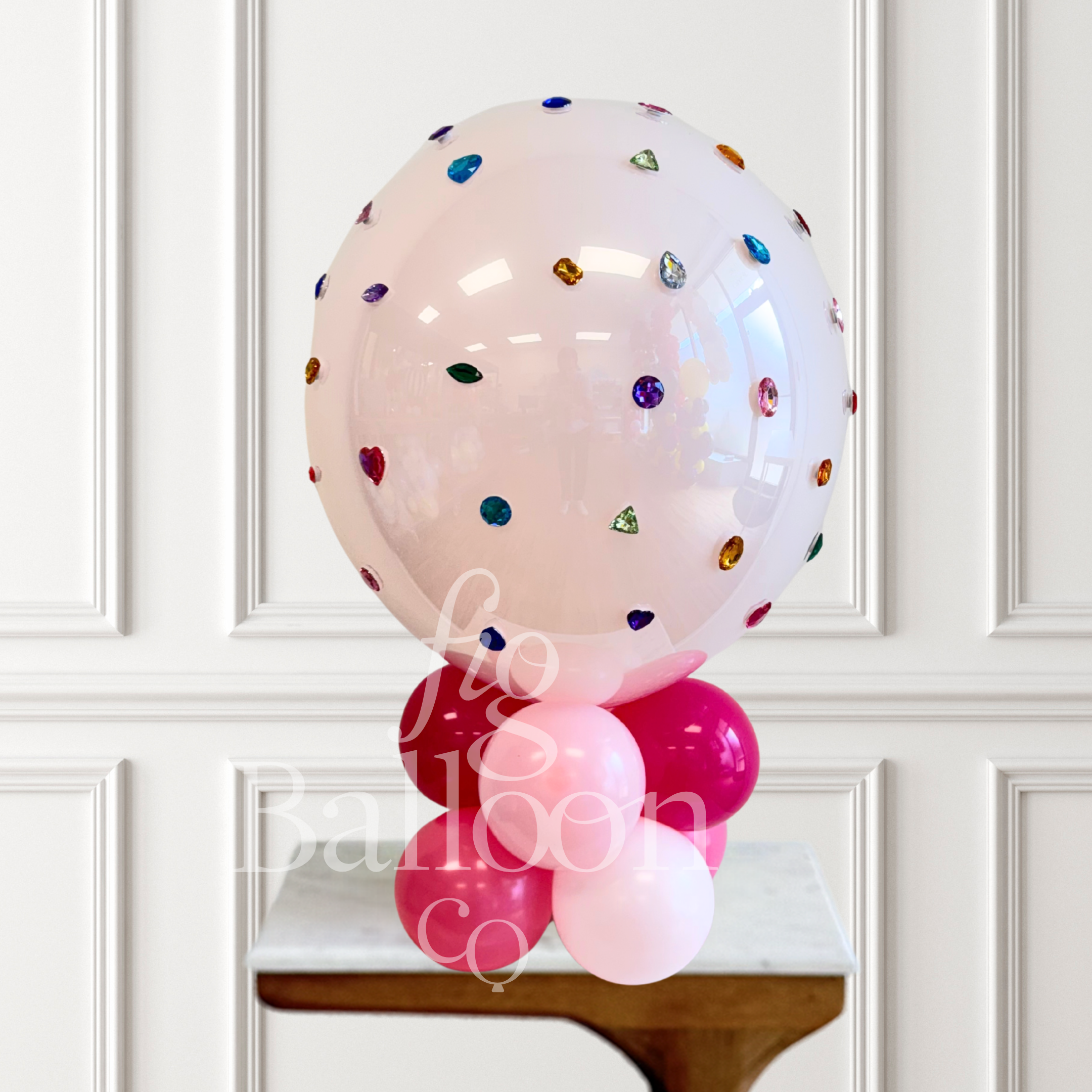 A large clear pink balloon decorated with scattered colorful gem-like confetti, displayed on top of a cluster of pink and light pink balloons on a small table against a white paneled wall.