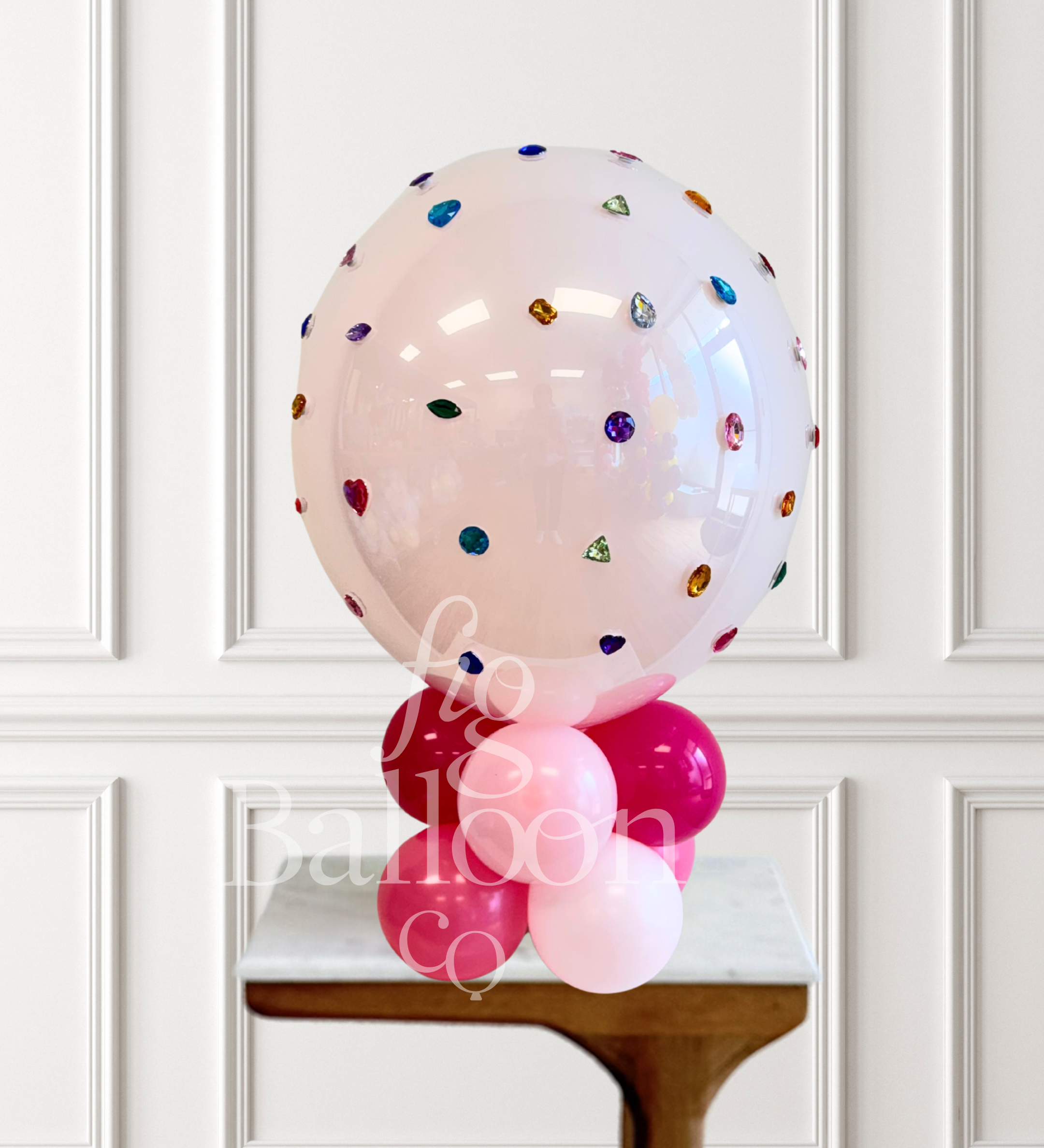 A large clear pink balloon decorated with scattered colorful gem-like confetti, displayed on top of a cluster of pink and light pink balloons on a small table against a white paneled wall.