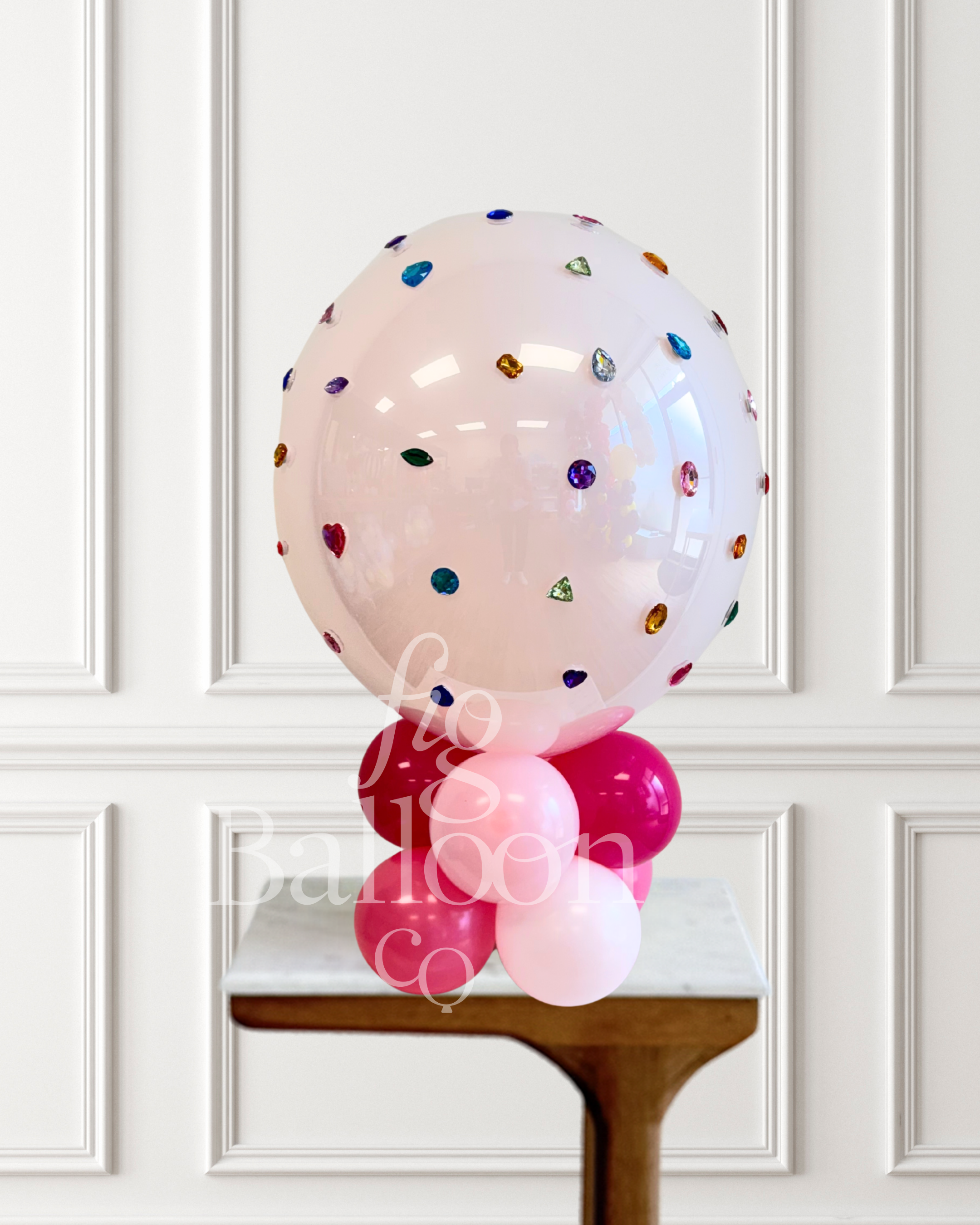 A large clear pink balloon decorated with scattered colorful gem-like confetti, displayed on top of a cluster of pink and light pink balloons on a small table against a white paneled wall.