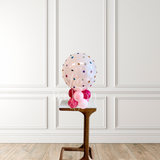 A clear round balloon filled with multicolored confetti stands on a small wooden pedestal in front of a white paneled wall and light wood floor.