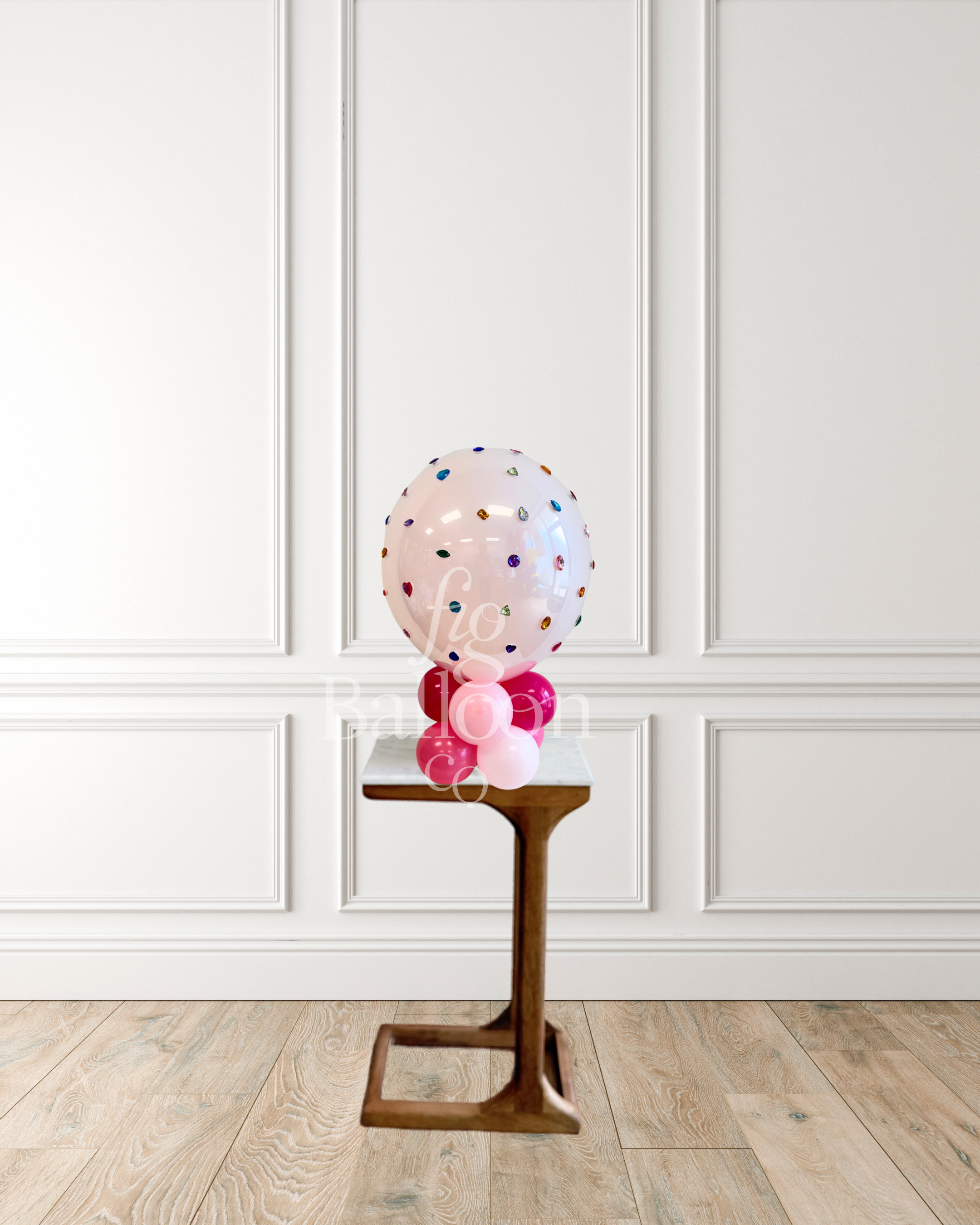 A clear round balloon filled with multicolored confetti stands on a small wooden pedestal in front of a white paneled wall and light wood floor.