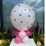 A large glossy white balloon decorated with scattered colorful jewel stickers, supported by pink and white balloons at the base, displayed on a marble pedestal in front of a window with decorative plants.