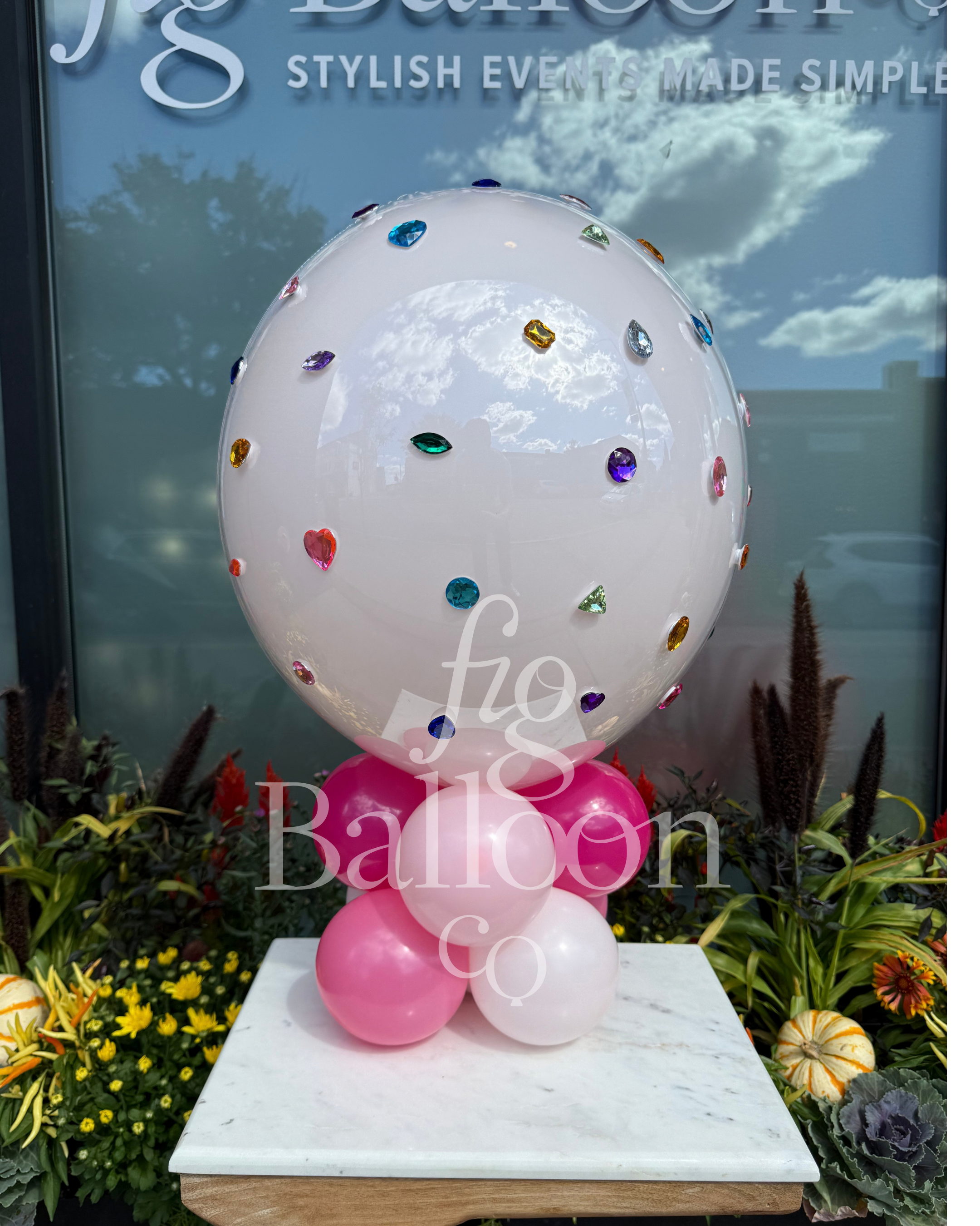 A large glossy white balloon decorated with scattered colorful jewel stickers, supported by pink and white balloons at the base, displayed on a marble pedestal in front of a window with decorative plants.