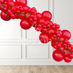 A red balloon garland made of glossy red latex balloons with small green leaf accents, displayed diagonally against a white paneled wall and light wood floor.