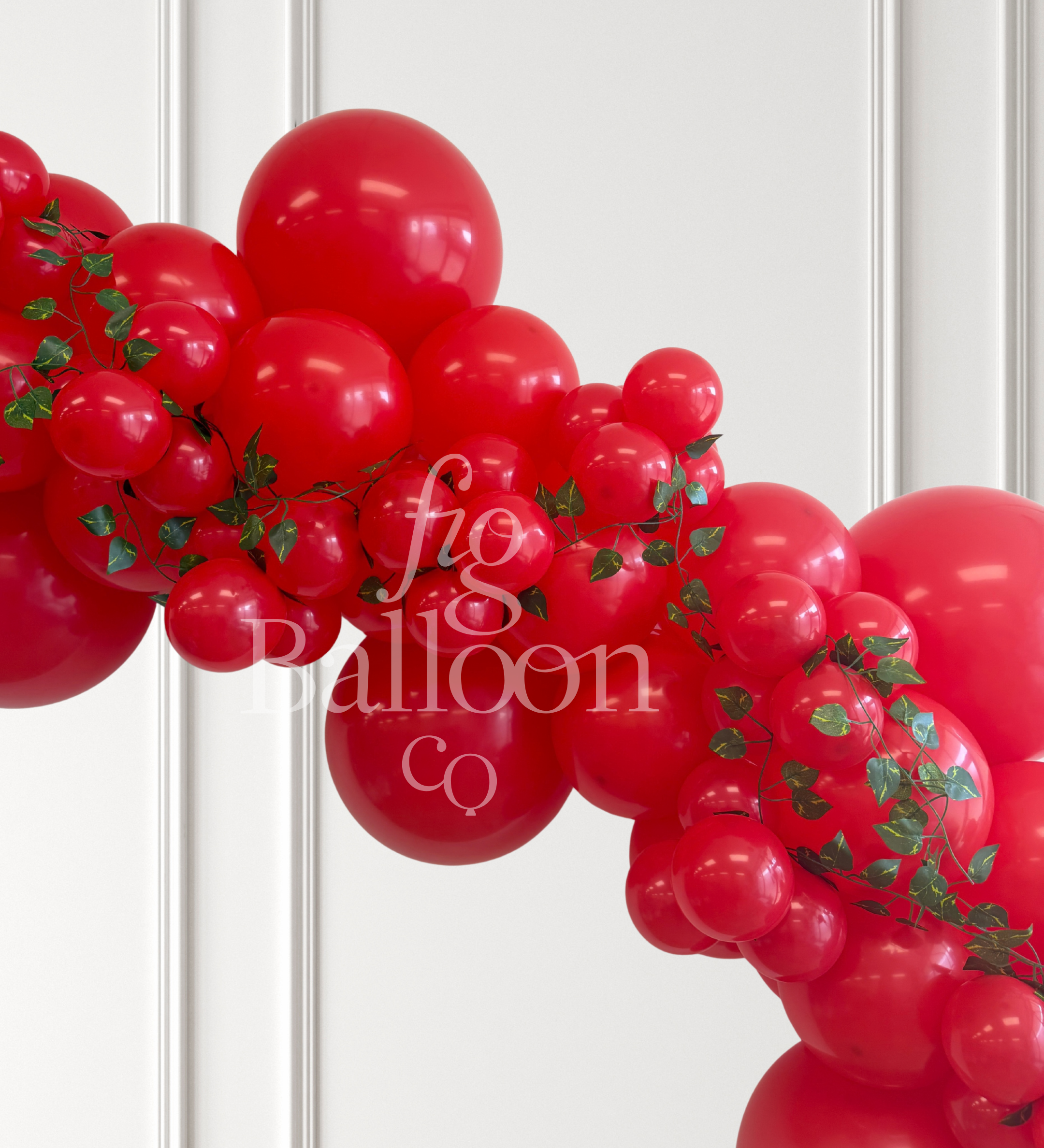 A red balloon garland arch decorated with green ivy vines, shown against a white paneled wall. A semi-transparent watermark reading 'fig balloon co' is overlaid across the center.