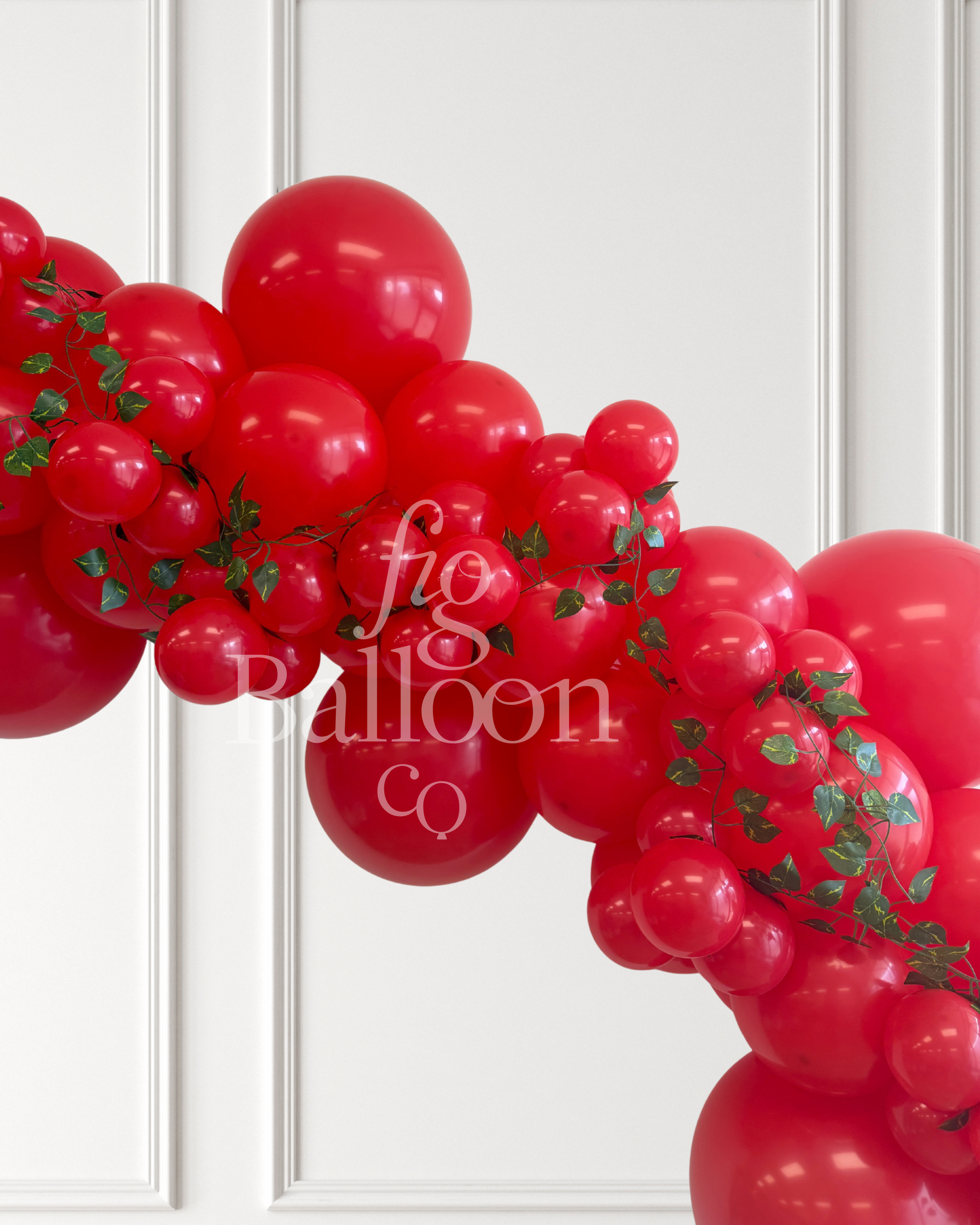 A red balloon garland arch decorated with green ivy vines, shown against a white paneled wall. A semi-transparent watermark reading 'fig balloon co' is overlaid across the center.