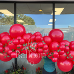 A large balloon garland arrangement in glossy red, decorated with small green leaf accents, displayed in front of a glass storefront window. A person in a teal shirt and dark pants is partially visible behind the balloon cluster.