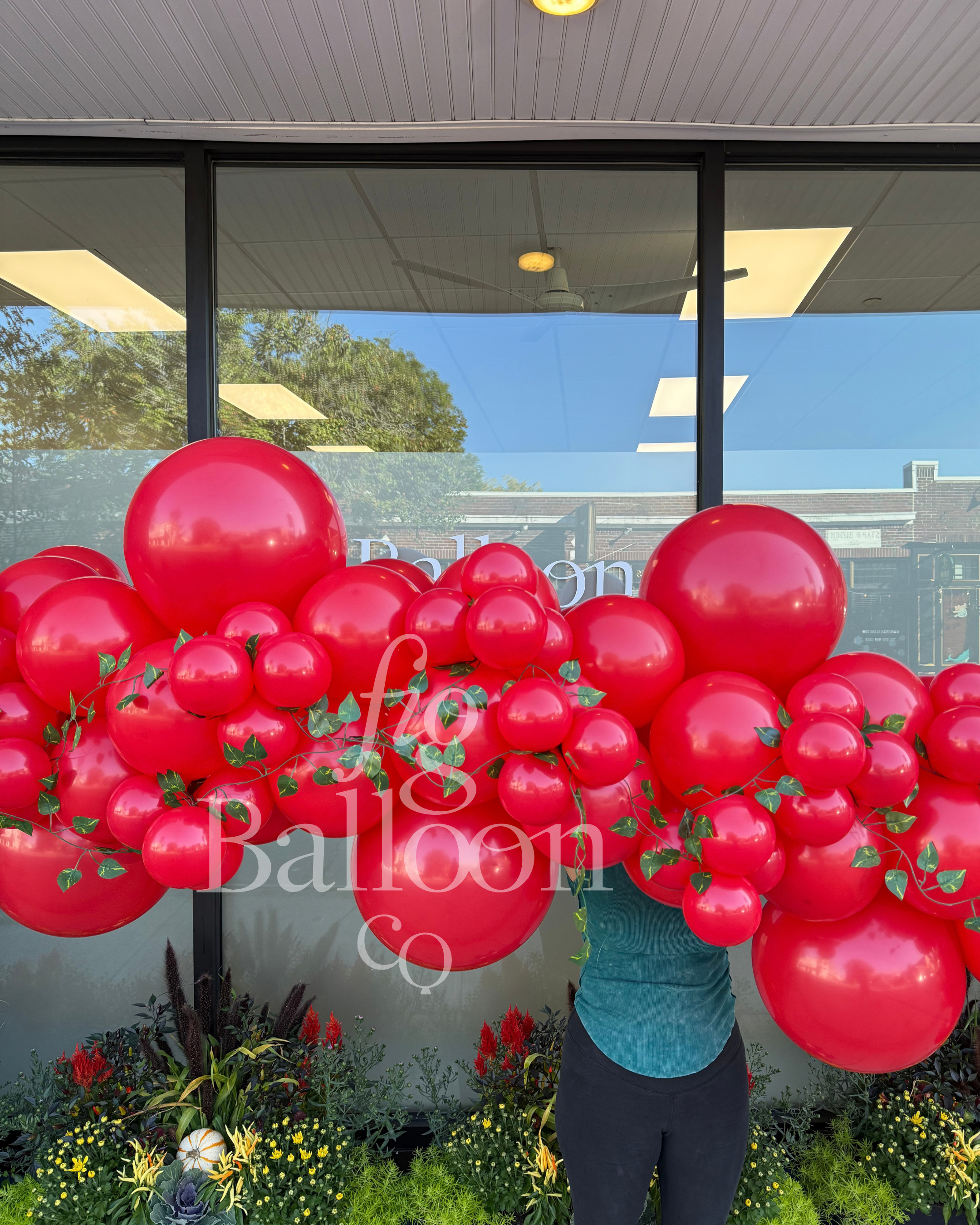A large balloon garland arrangement in glossy red, decorated with small green leaf accents, displayed in front of a glass storefront window. A person in a teal shirt and dark pants is partially visible behind the balloon cluster.