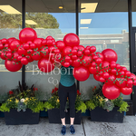 A person stands in front of a storefront window holding a large balloon garland made of many glossy red balloons with small green leaf accents. Planter boxes sit along the window below.