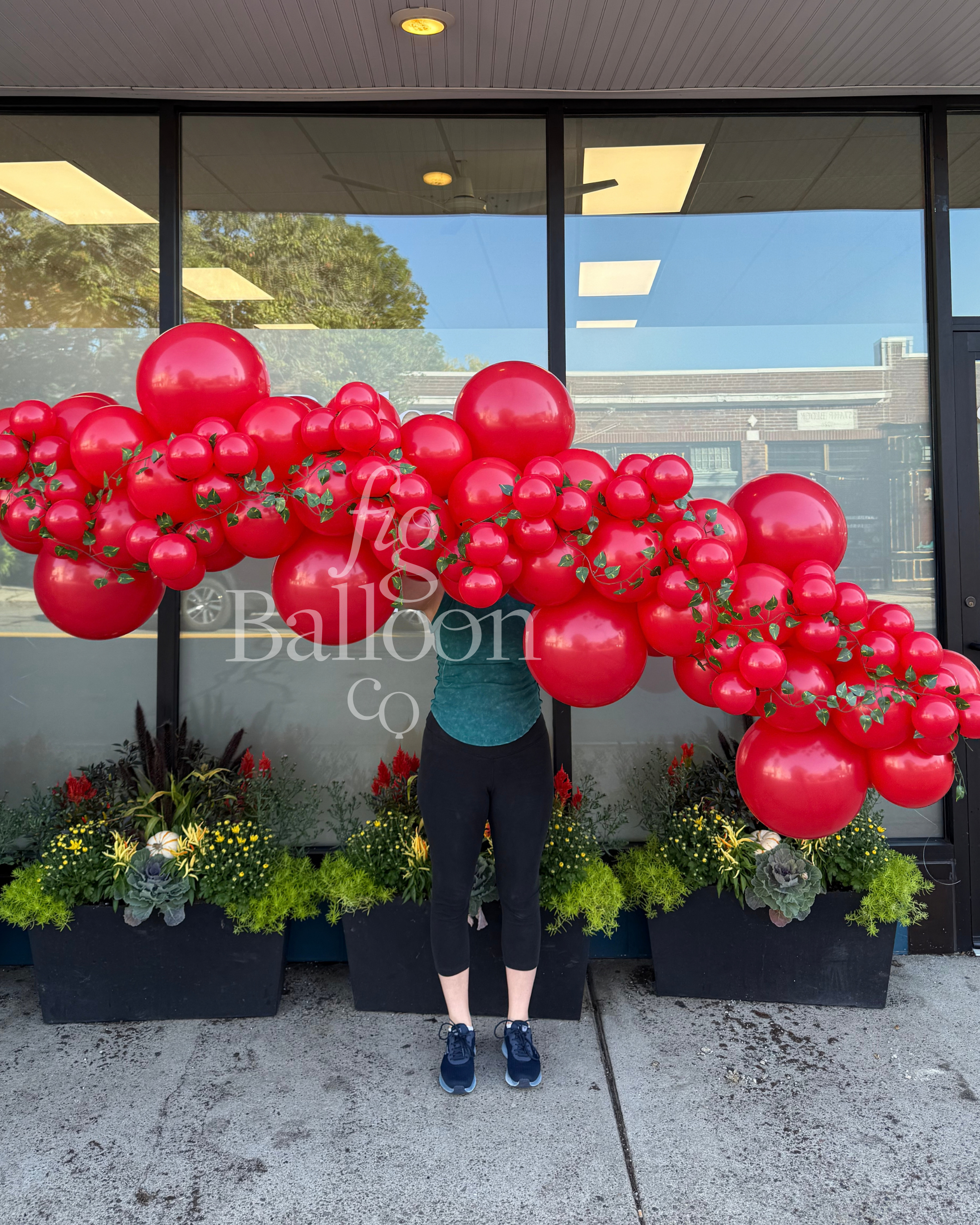 A person stands in front of a storefront window holding a large balloon garland made of many glossy red balloons with small green leaf accents. Planter boxes sit along the window below.